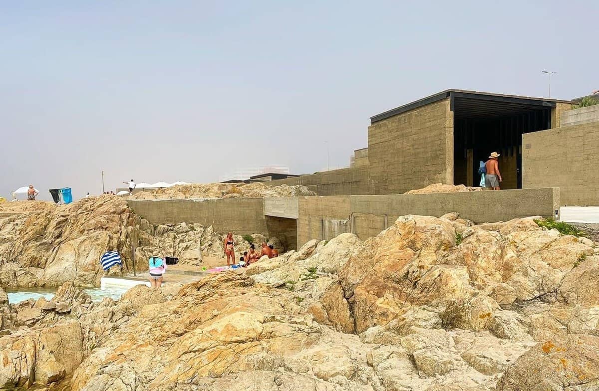 Rocky coastal bathing area with concrete modernist structures built into the landscape, with people swimming, sunbathing and exploring the rocks on a sunny day.